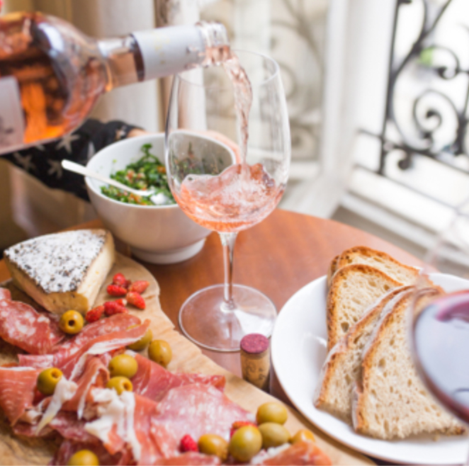 A bottle of rose being poured into a glass on a table with a charcuterie board next to it